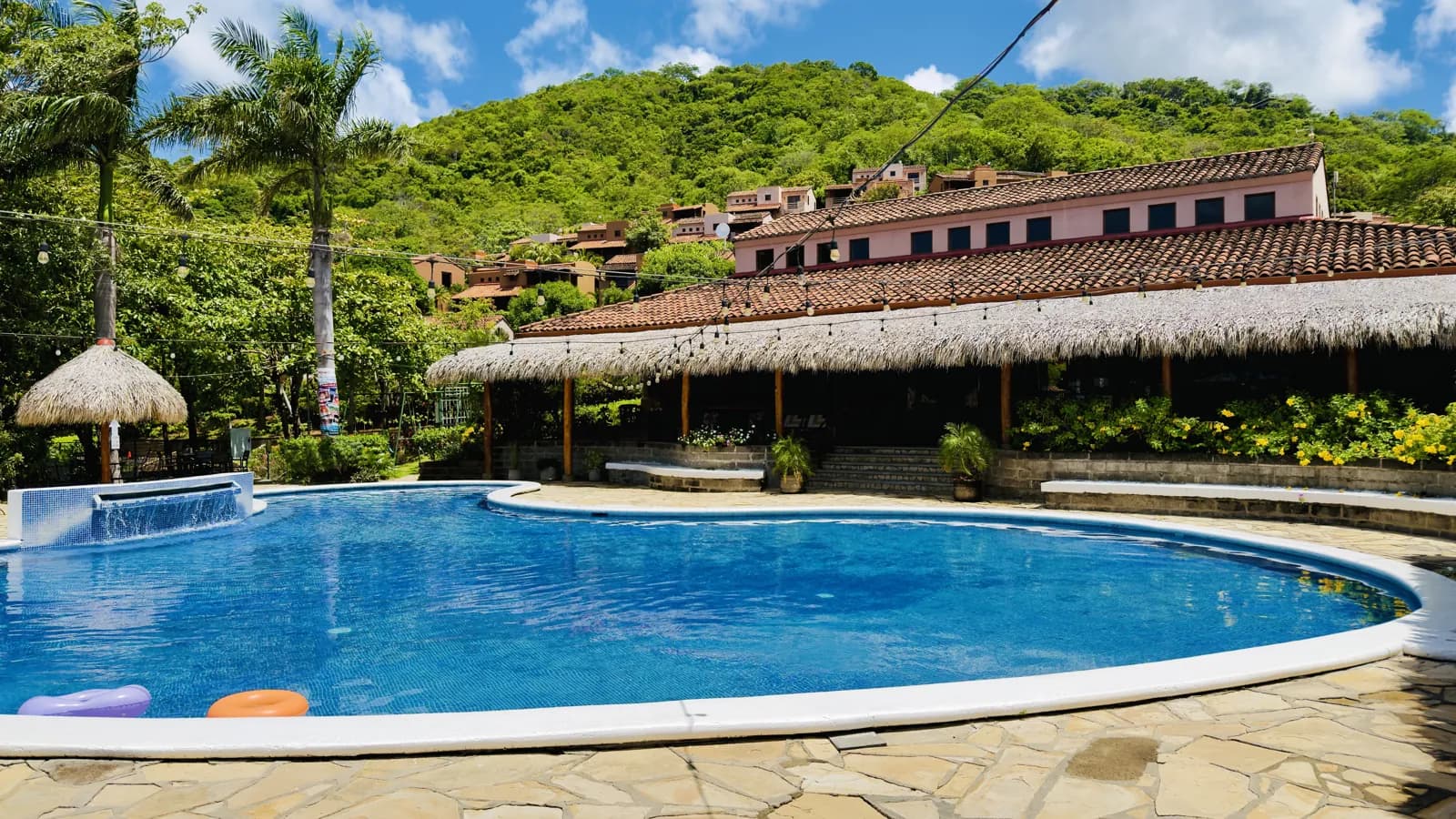 Terraza de Palermo under its palapa roof beside the pool, with the villas on the hill behind